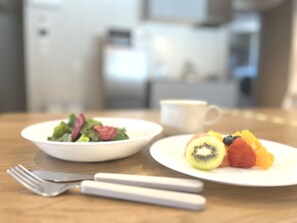 Una mesa de comedor con un tazón blanco de ensalada, un plato con fruta, una taza blanca y un tenedor.