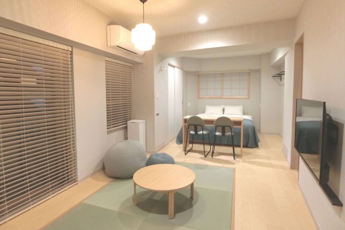A modern, compact room with a bed, a dining table with chairs, a small round table, a television, and a window with blinds.