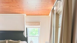 Une chambre avec un plafond en bois, un lit doté d’une tête de lit sombre, une table de chevet avec une lampe et une fenêtre avec des rideaux.