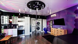 Una cocina moderna con armarios blancos, un microondas y una estufa. Hay una mesa de comedor con sillas y un sofá negro. La habitación tiene una decoración en tonos negros y morados, con un ventilador de techo y un televisor montado en la pared.