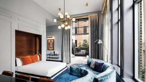 A modern hotel room with a large bed, a curved blue sofa, a dining area, and a view of the outdoors through large windows. - 1-Bedroom Suite at a 4⭐️ Hotel (Nashville)