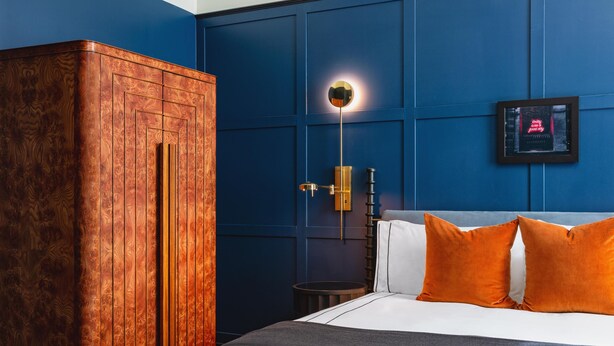 Un dormitorio con un armario de madera, una cama con almohadas blancas y anaranjadas, una mesita de noche y un aplique de pared.