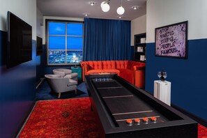 A room with a shuffleboard table, a red sofa, a grey armchair, a blue curtain, and a city view through a window.