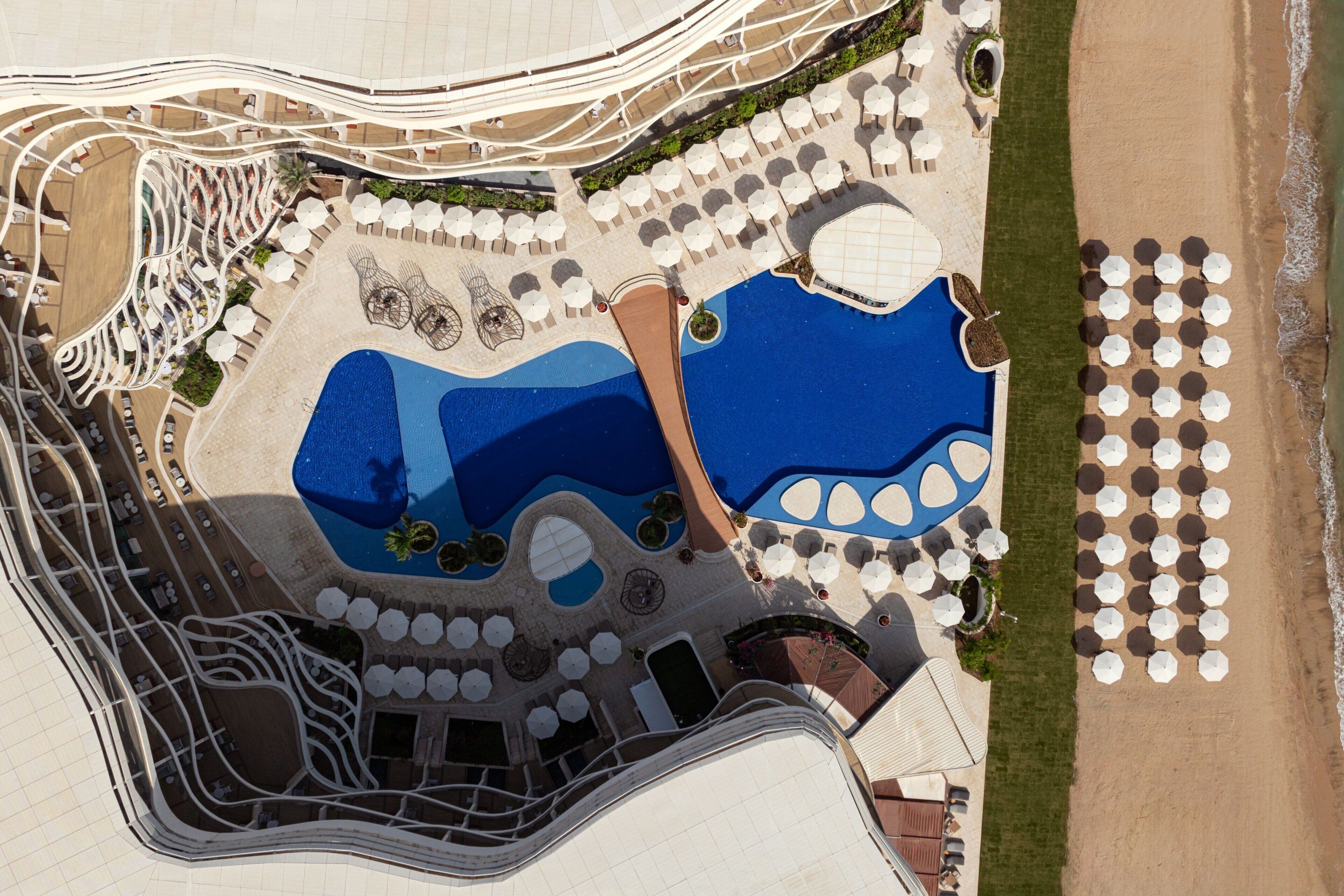 An aerial view of a resort with a large pool, multiple lounge areas, and a beach area with umbrellas.