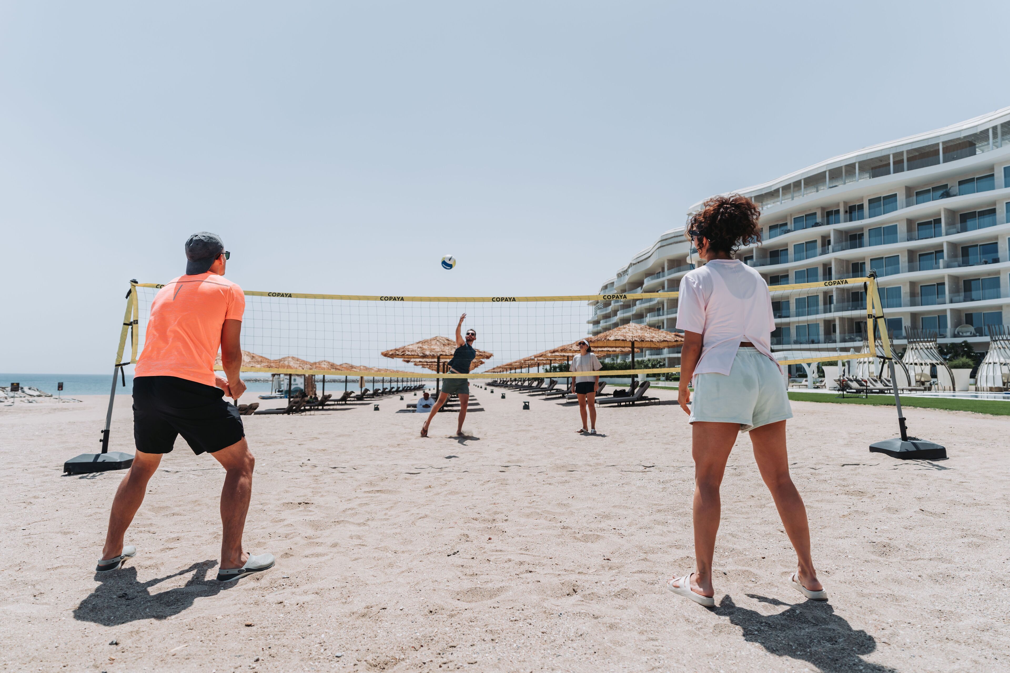 Un match de volleyball de plage avec un filet portant l’inscription « COPAYA » et, en arrière-plan, un bâtiment doté de balcons.