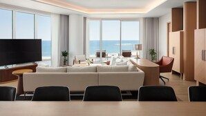 A modern hotel room with a large sofa, a dining table, and a TV. There are large windows with a view of the sea. - The St. Regis Al Mouj Muscat Resort (Muscat)