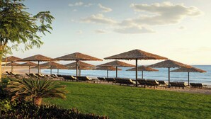 A beach with rows of sun loungers and thatched umbrellas, a palm tree, and a clear view of the sea. - The St. Regis Al Mouj Muscat Resort (Muscat)