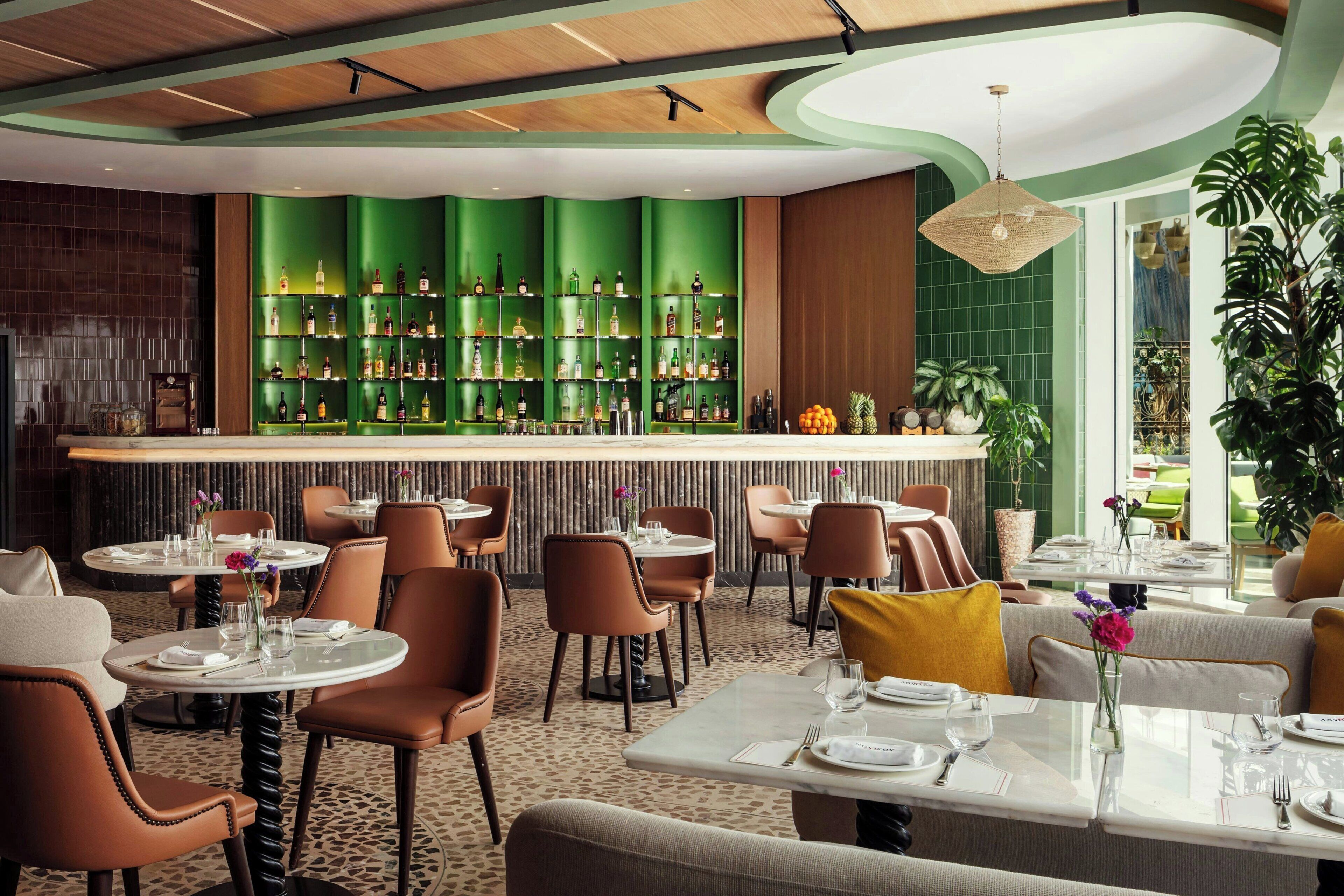 A restaurant with a green bar, round tables, and brown chairs.