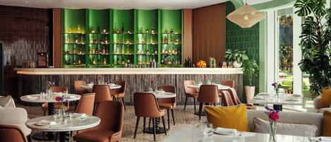 A restaurant with a green bar, round tables, and brown chairs.