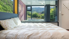 Un dormitorio con un ventanal grande, una cama con ropa de cama blanca y vistas a un paisaje verde.