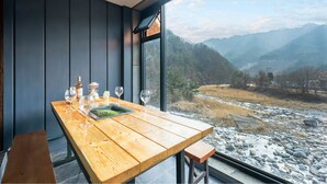 Une table en bois, ornée de verres à vin et d’une bouteille, est placée devant une fenêtre donnant sur un paysage montagneux.