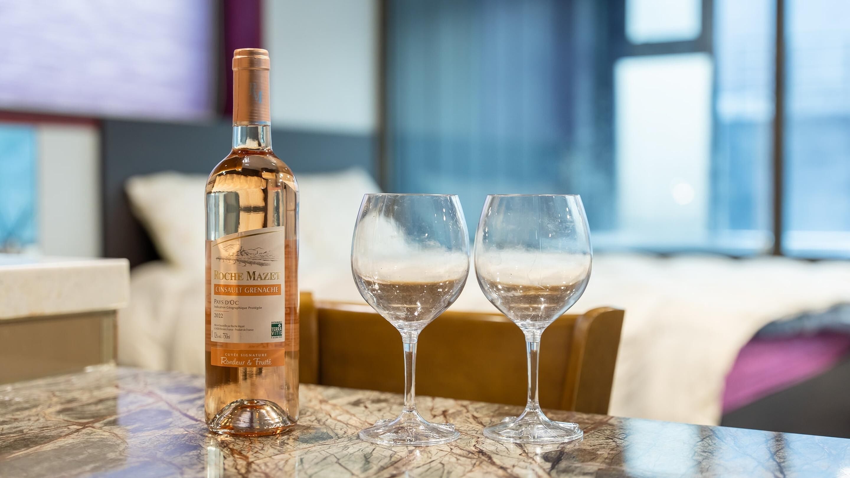 A bottle of Cinsault Grenache wine and two empty wine glasses on a marble table in a hotel room.