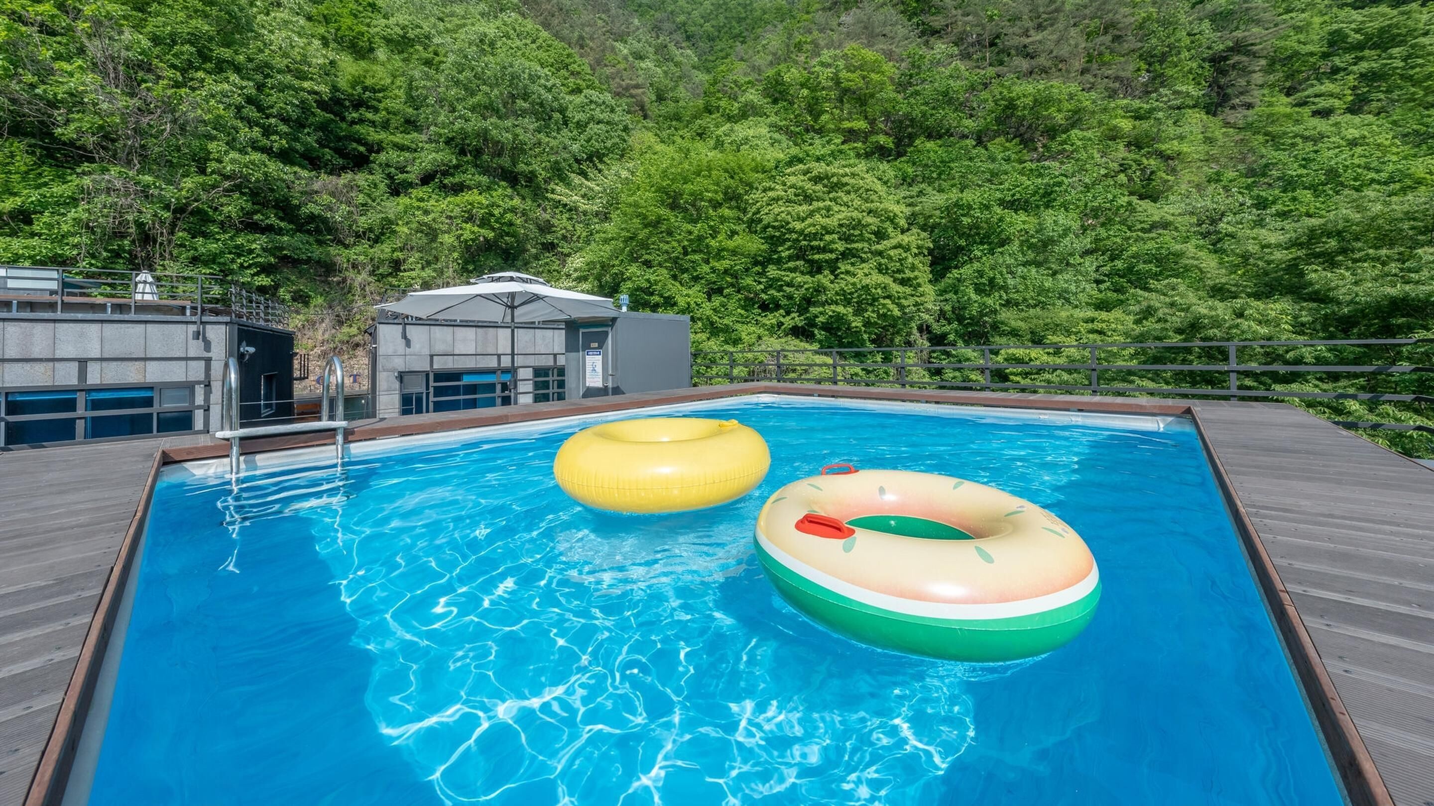 Ein Schwimmbecken mit zwei aufblasbaren Ringen, umgeben von einer Holzterrasse und einem Gebäude mit Glasfassade.