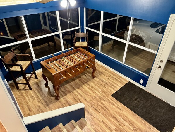 A room with a wooden foosball table, a chair, and a window with a view of a car outside.
