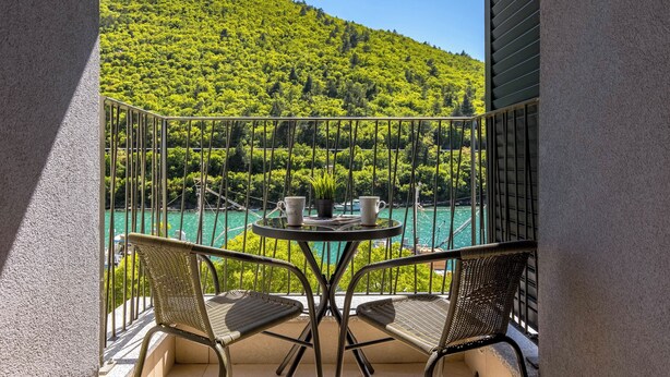 Un balcon agrémenté de deux fauteuils en osier et d’une petite table ronde, offrant une vue sur une mer turquoise et une colline verdoyante.
