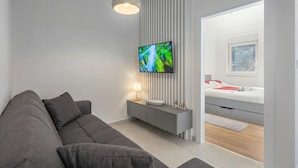 A modern living room with a grey sofa, a flat-screen TV mounted on a wall, a bedside table with a lamp, and a bed with a white headboard. - Sea-View Balcony Apt Lady Laura, Plomin, Croatia (Plomin)