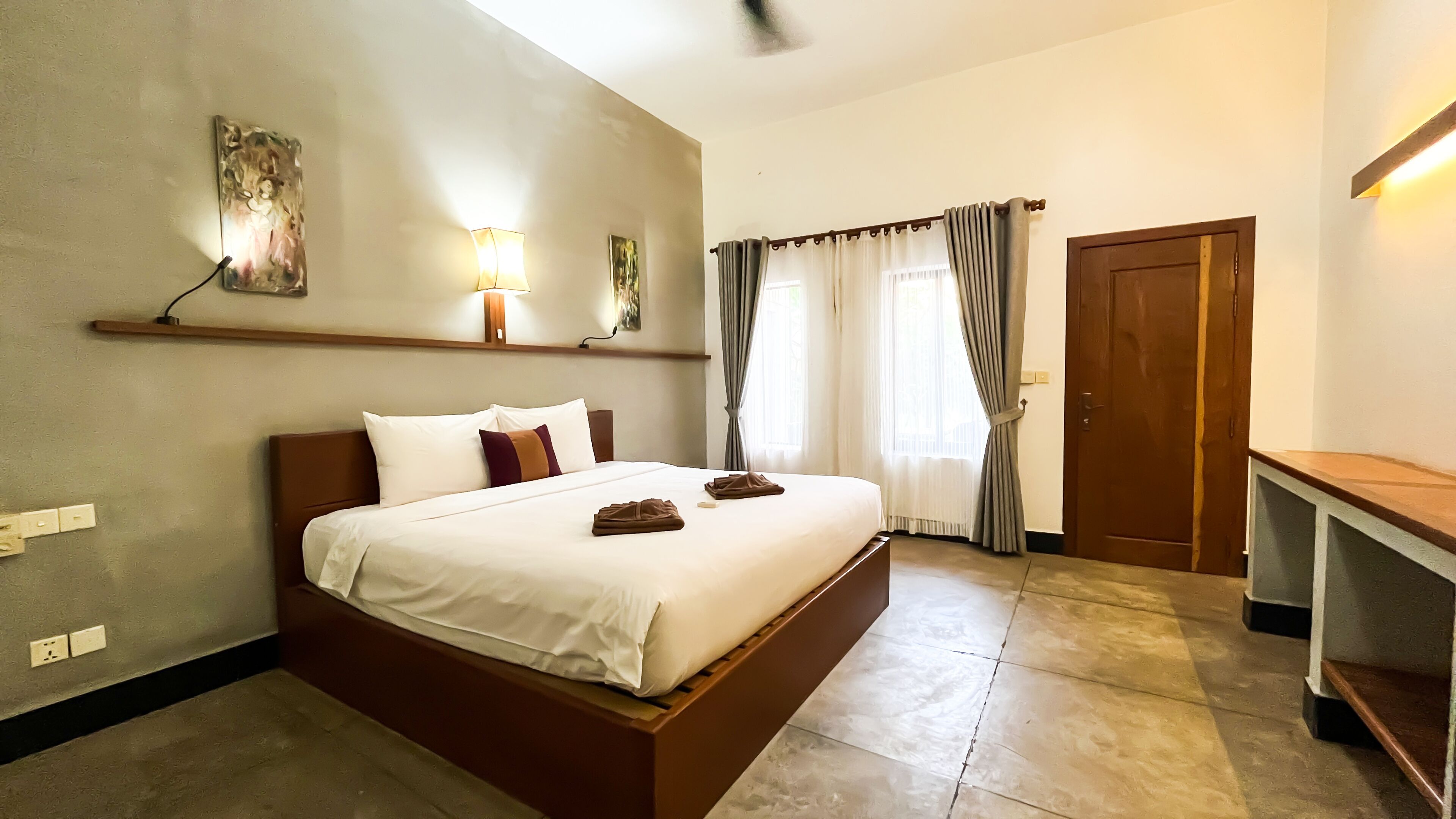 A neatly made bed with white linens and two brown towels, a bedside table, a wall-mounted lamp, and a door with a curtain.