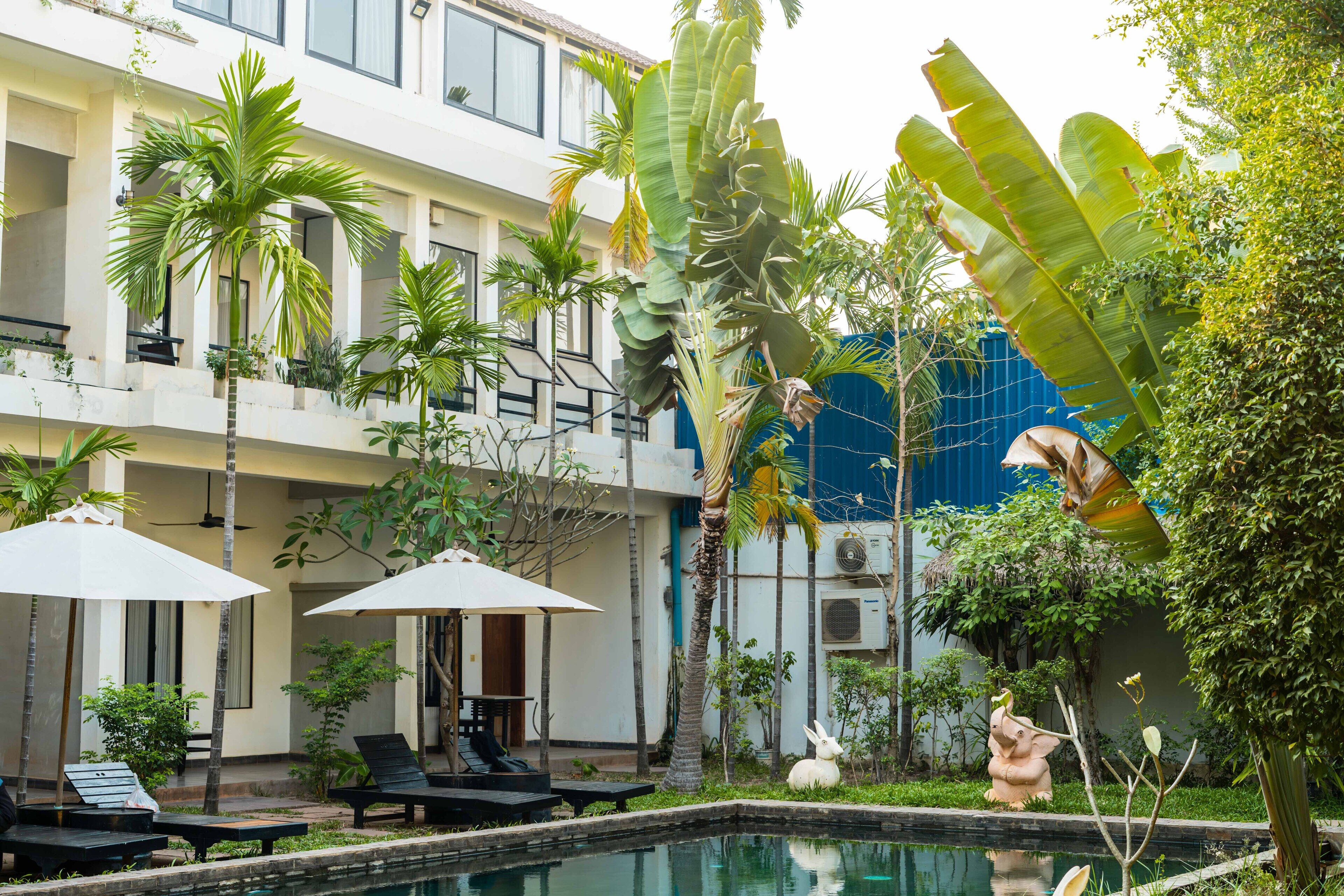 Un área junto a la piscina con sillones de descanso y sombrillas, rodeada de plantas tropicales y una estatua.
