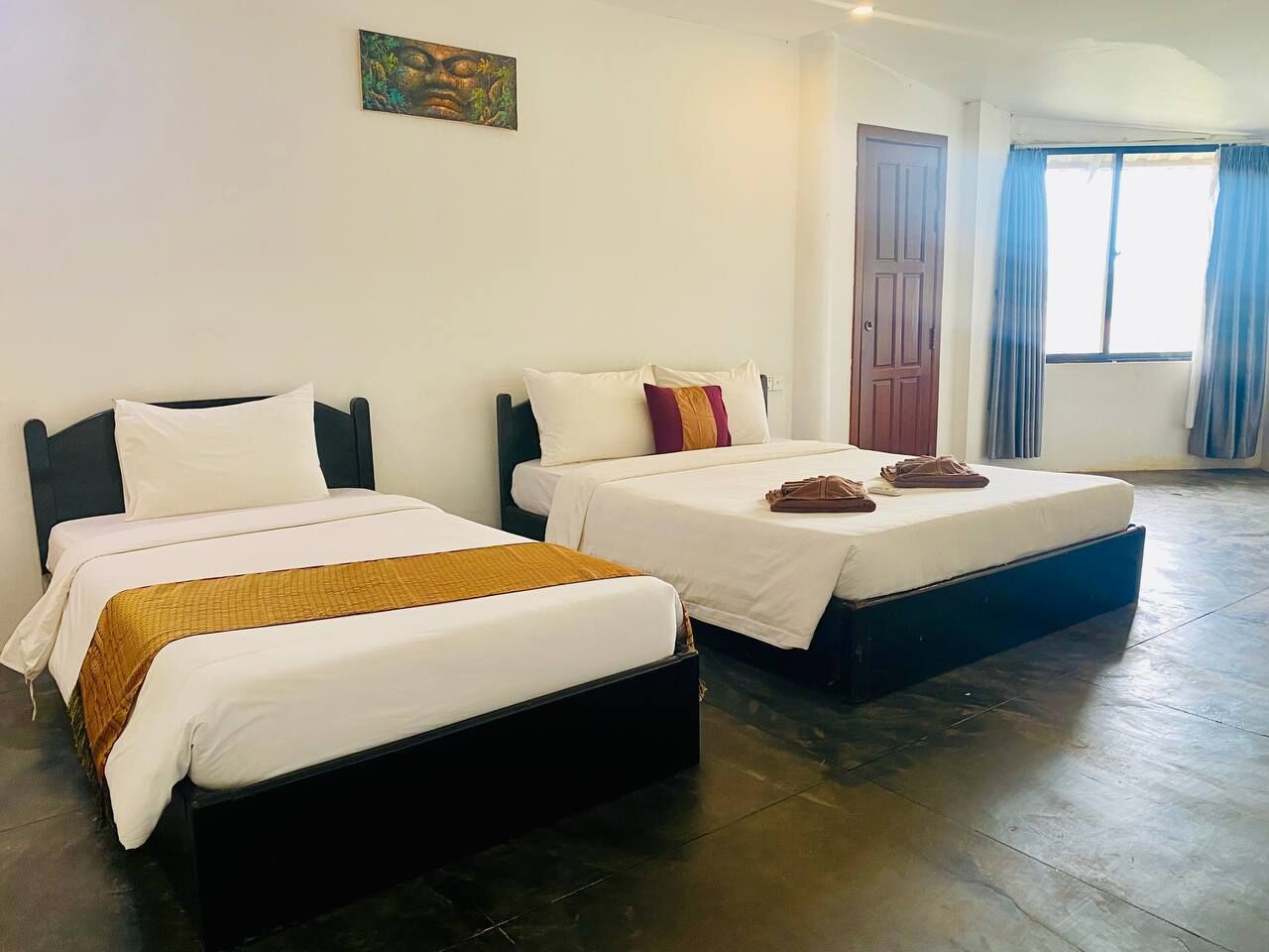 Two single beds with dark frames and white bedding, a wooden floor, a door, and a window with blue curtains.