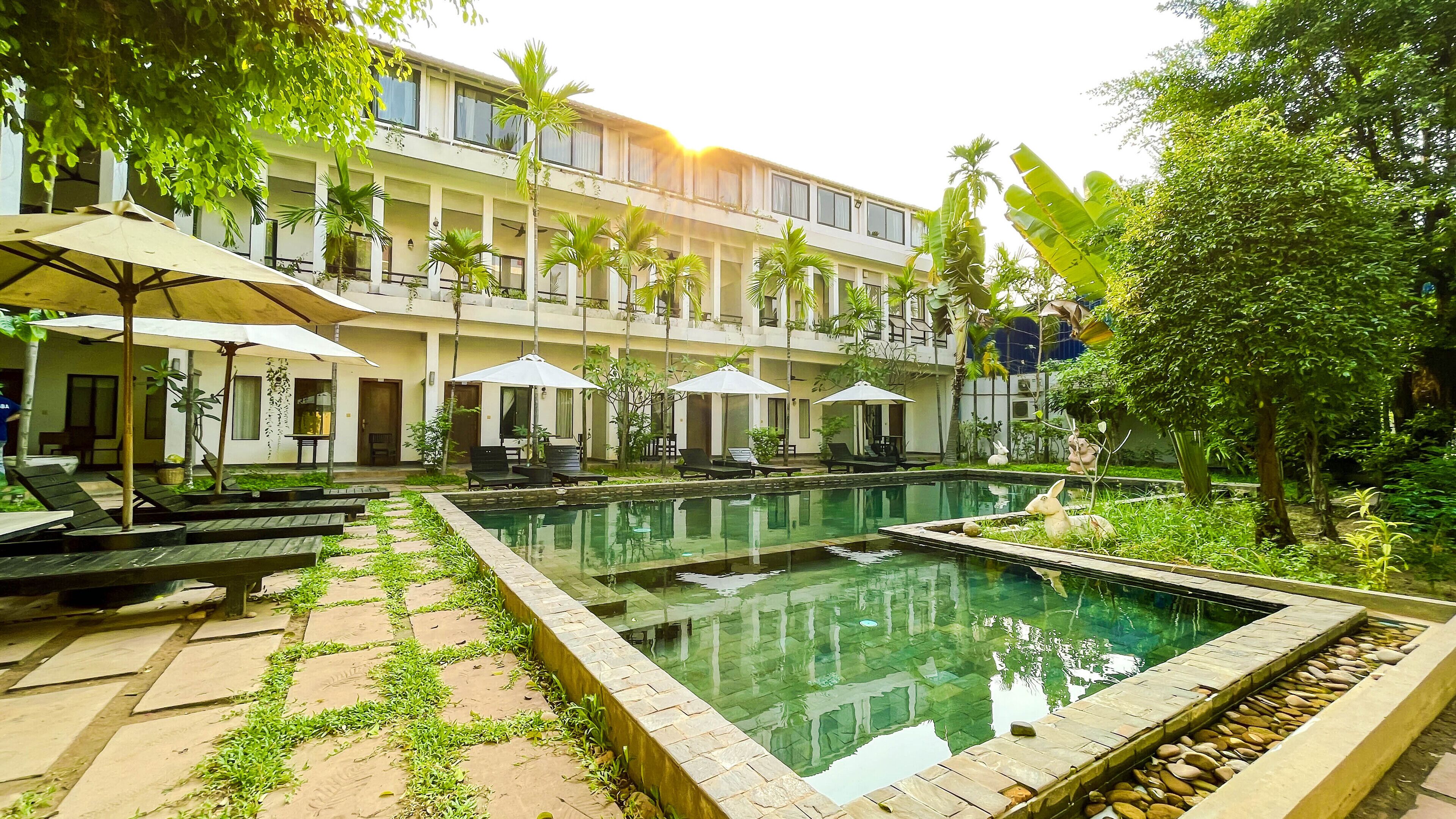 A pool area with lounge chairs and umbrellas, surrounded by greenery and a multi-story building.