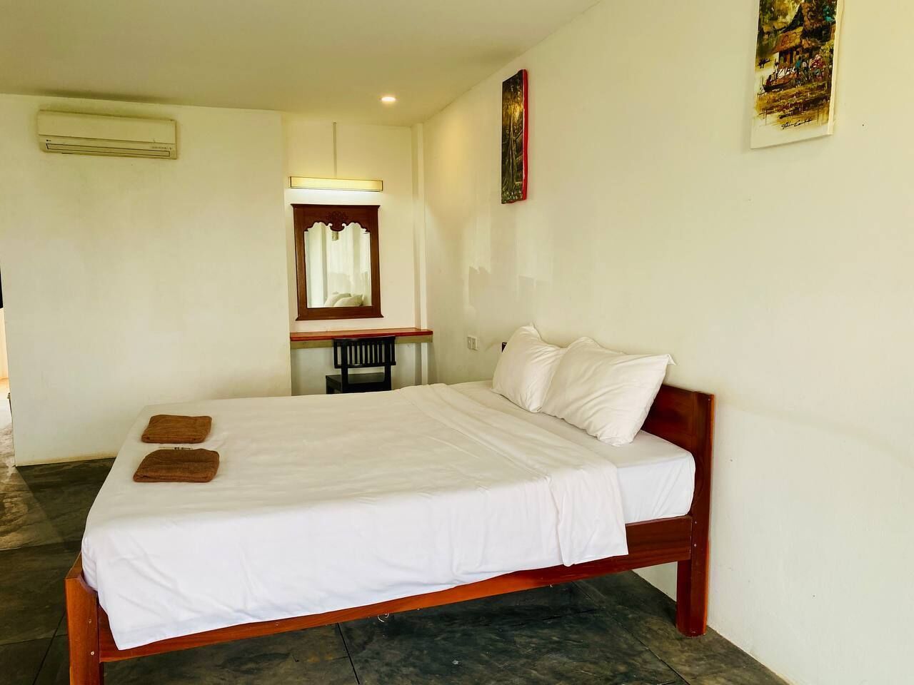 A single bed with white bedding and two brown towels, a wooden headboard, a small table with a chair, and a wall-mounted air conditioner.