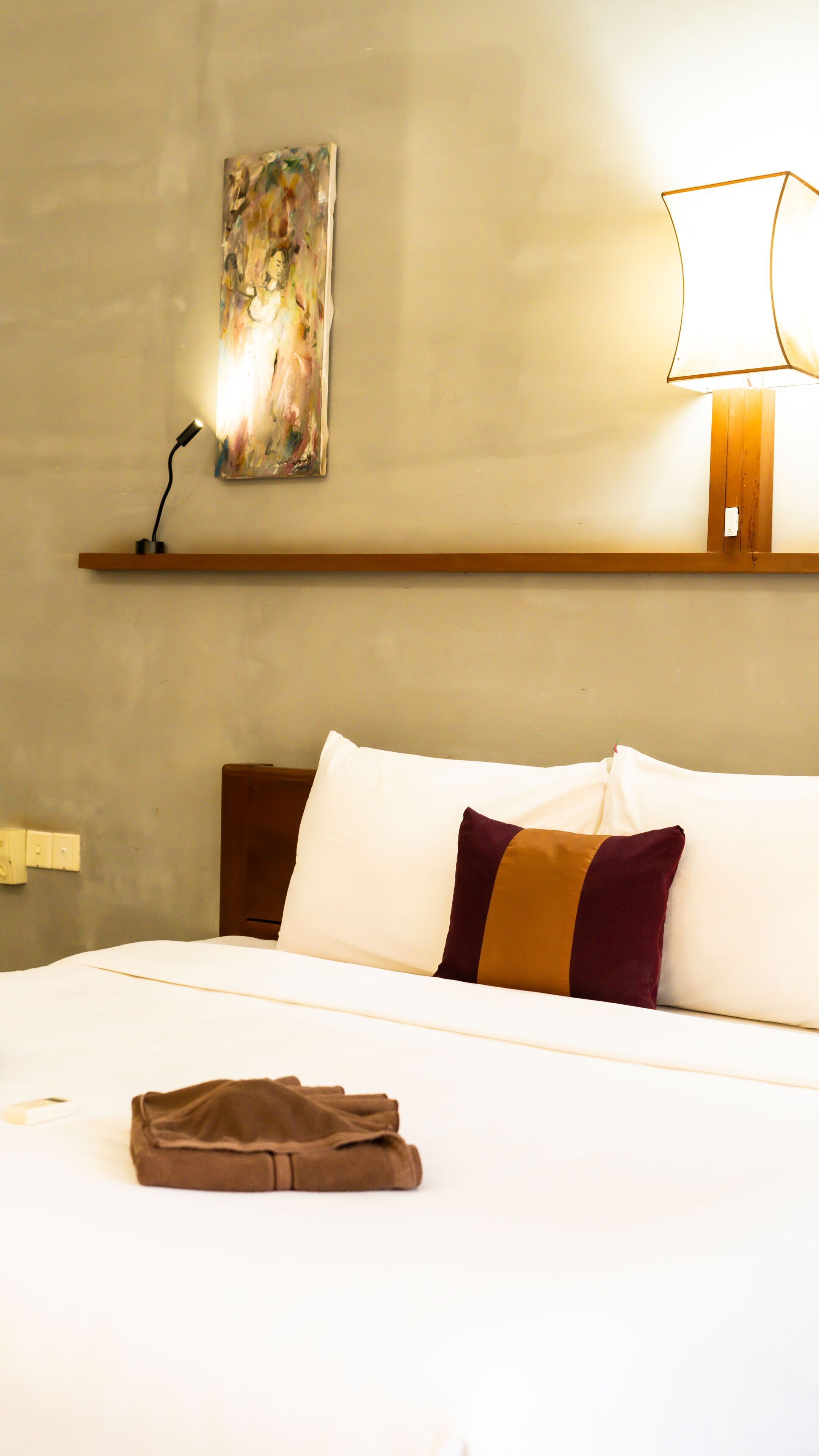 A hotel room with a neatly made bed, a painting on the wall, and a lamp on a bedside table.