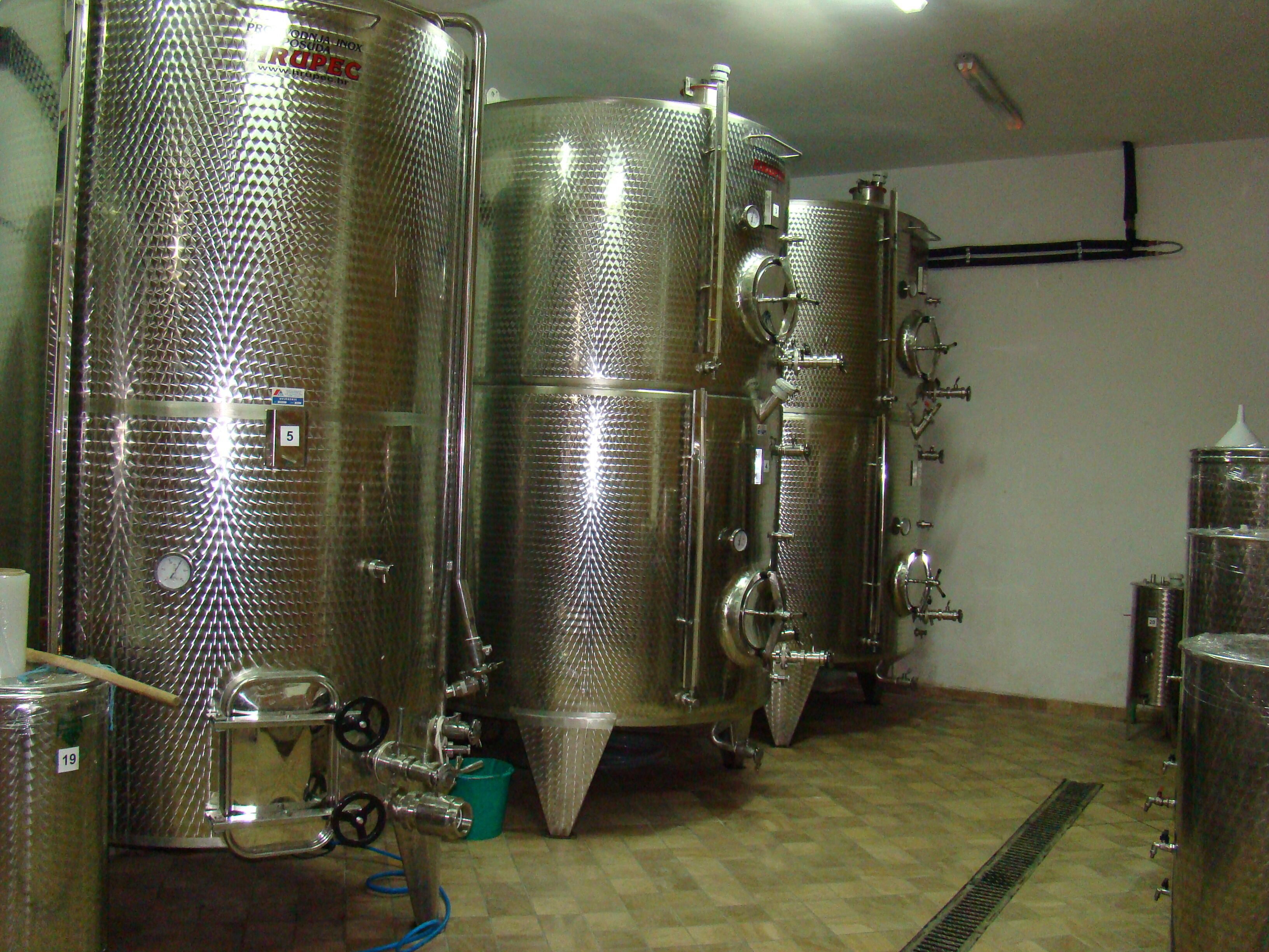 Stainless steel fermentation tanks in a brewery with a tiled floor and a vent system on the wall.