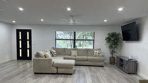 Un salon moderne avec un canapé d’angle, un ventilateur de plafond, une grande fenêtre avec des stores, un téléviseur fixé au mur et un plancher en bois.