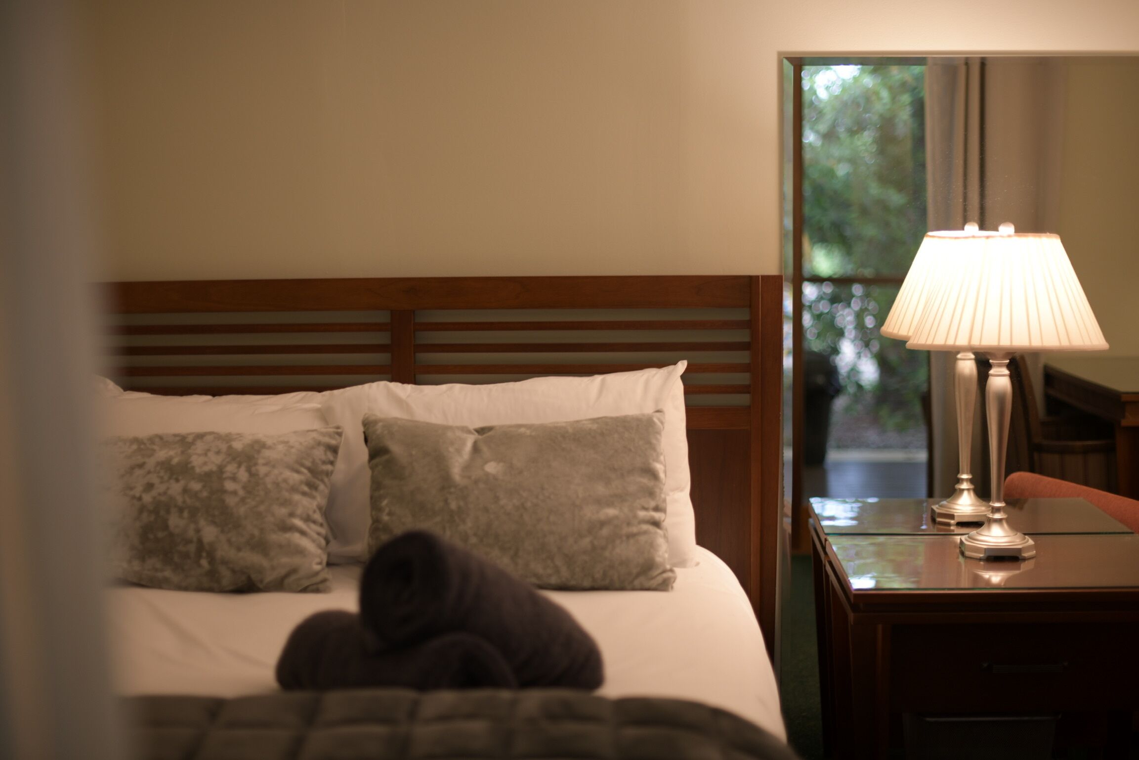 Habitación de hotel con cama, almohadas, mesita de noche con lámpara y ventana con vistas a zonas verdes.