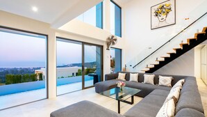 A modern living room with a large gray sectional sofa, a glass coffee table, and a view of a body of water through large sliding glass doors. - Luxury 5 Bedroom Modern Villa - KBR17 (Koh Samui)