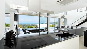 A modern kitchen with a large island, a dining area with a view, and a staircase leading to a balcony. - Luxury 5 Bedroom Modern Villa - KBR17 (Koh Samui)