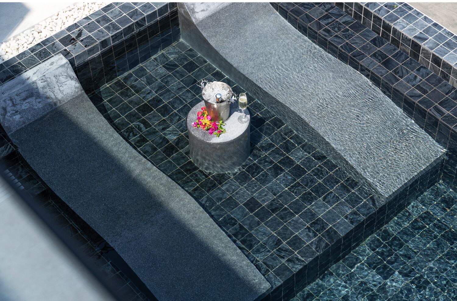 Un jacuzzi con borde de azulejos y un pedestal de concreto en el centro, que contiene un balde con hielo y una copa de champán.