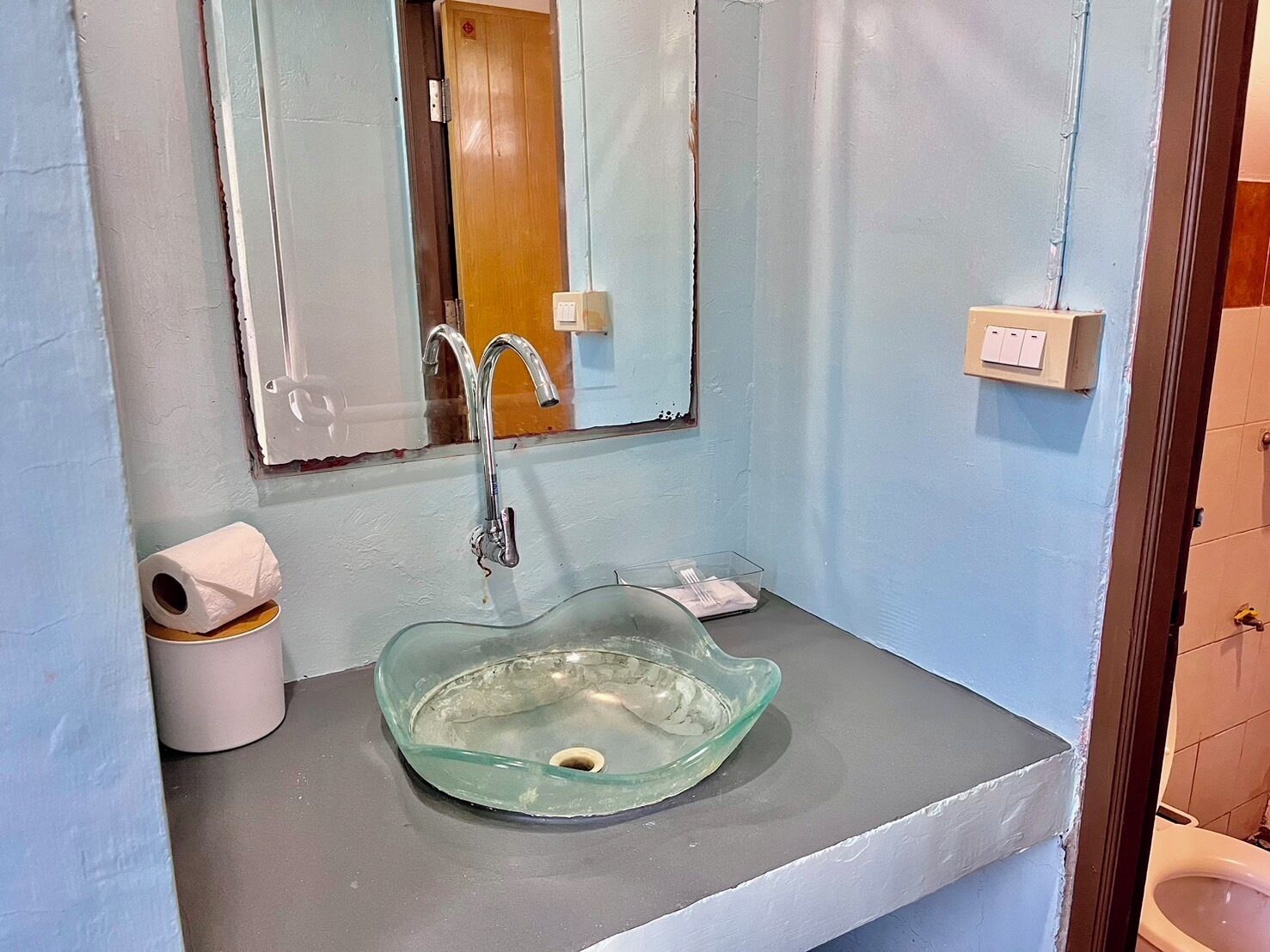 A bathroom with a glass sink, a mirror, a toilet, and a roll of toilet paper.