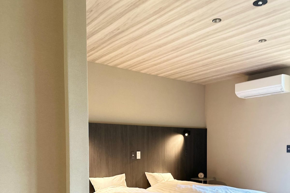 Une chambre d’hôtel moderne avec un parquet en bois disposé en chevrons, un grand lit avec du linge de lit blanc, une tête de lit sombre et un éclairage encastré au plafond.