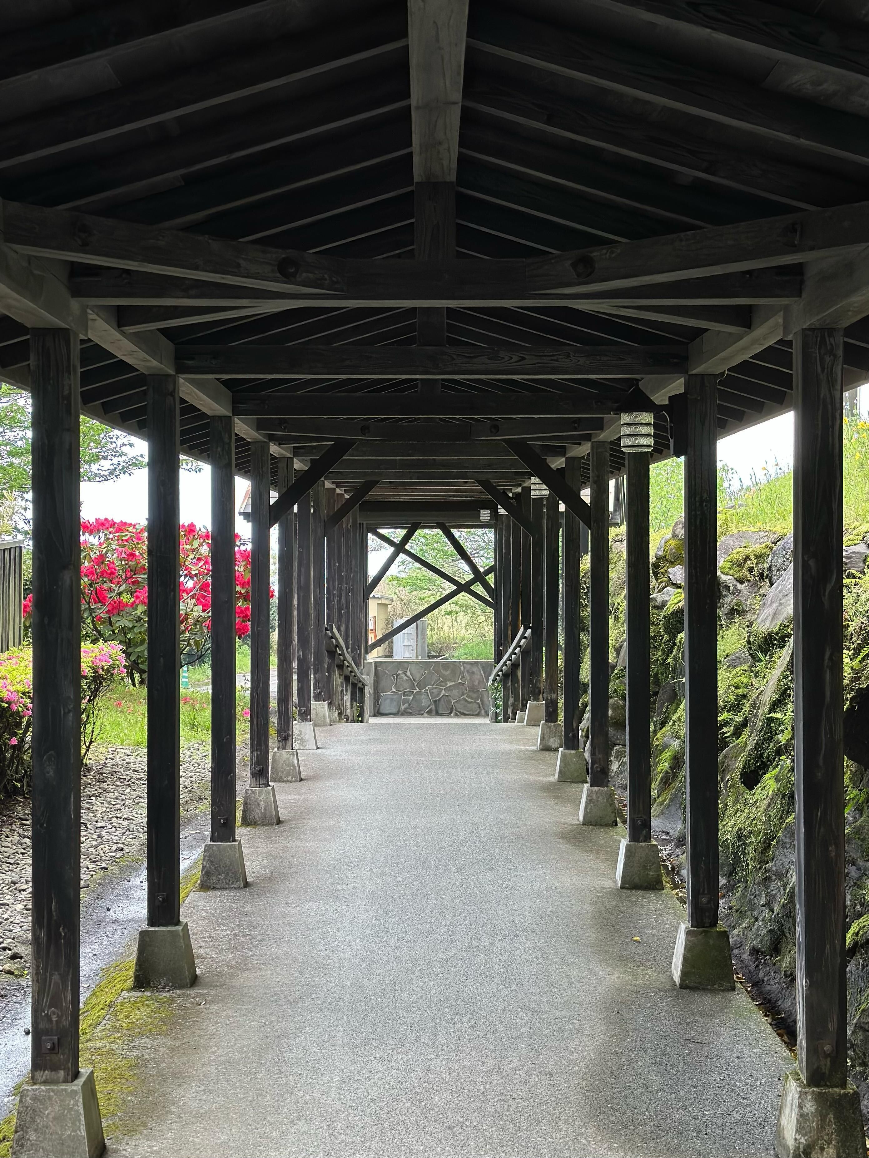 A covered walkway with wooden beams and stone pillars, leading to a stone wall.