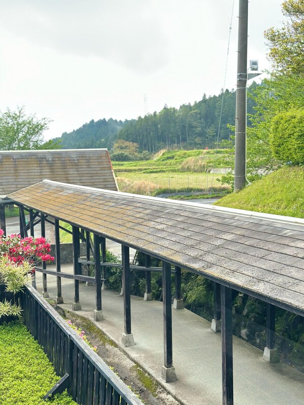 Un pasaje peatonal cubierto con techo de madera, sostenido por pilares negros, junto a un área ajardinada con flores y arbustos.