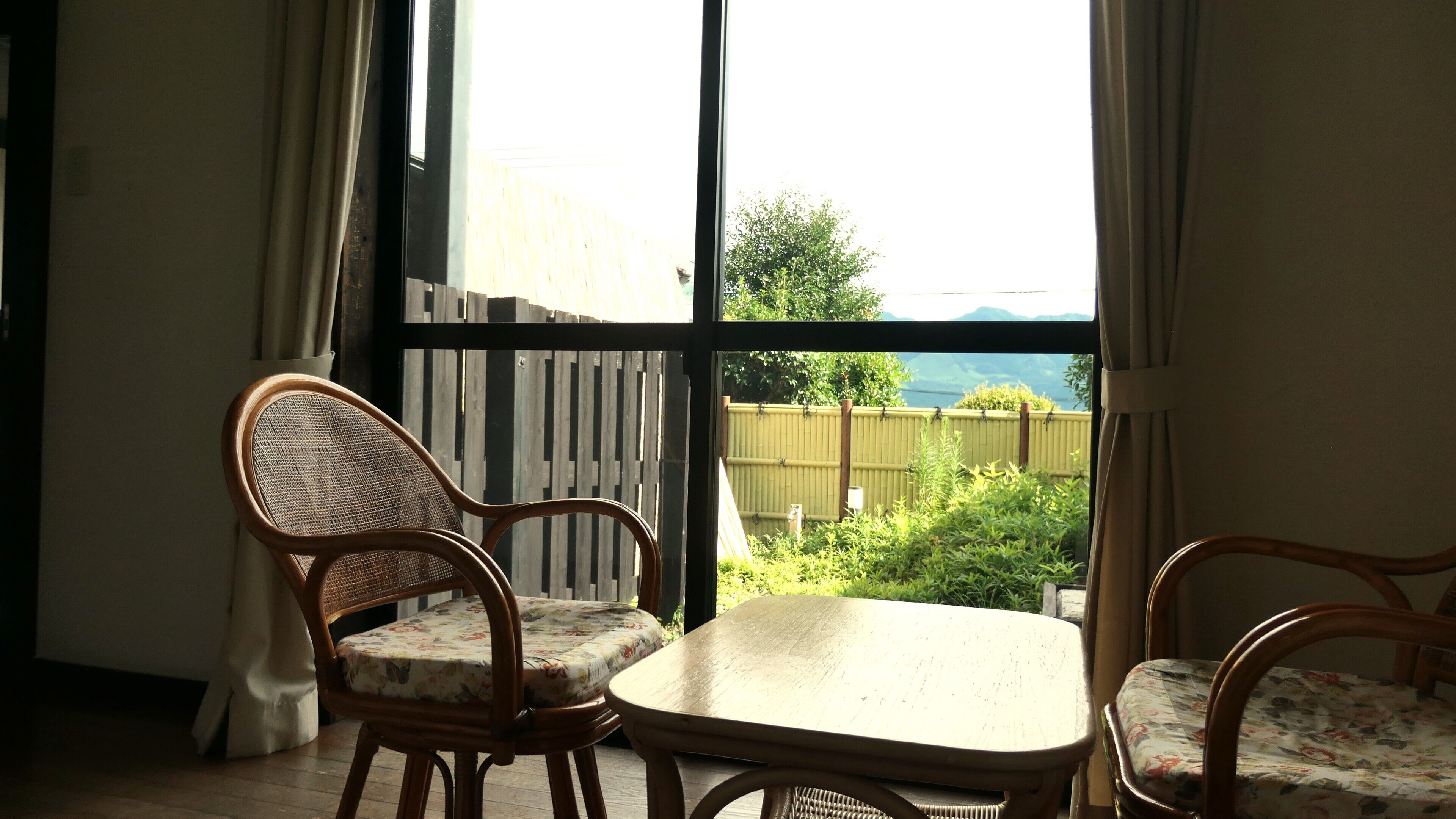 A room with a large window, wicker chairs, a small table, and a view of greenery and mountains.
