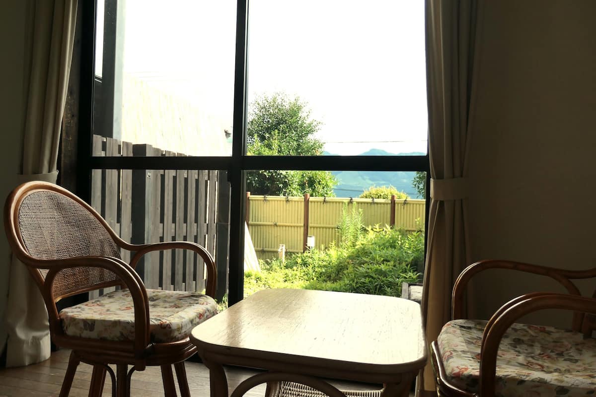 Une pièce dotée d’une grande fenêtre, de fauteuils en osier, d’une petite table et offrant une vue sur la verdure et les montagnes.