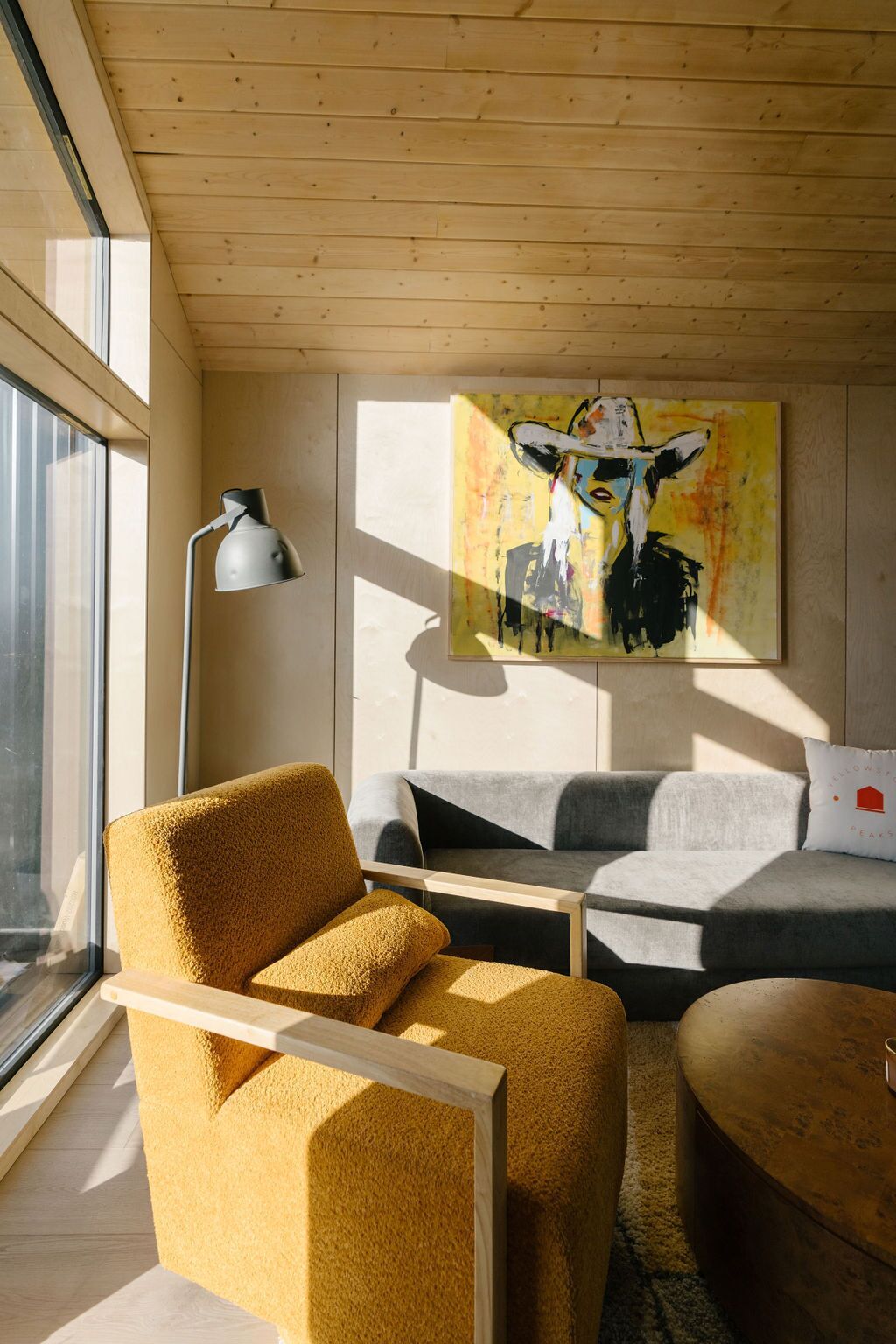 A modern living room with a yellow armchair, a grey sofa, and a round coffee table. There's a painting on the wall and a floor lamp.