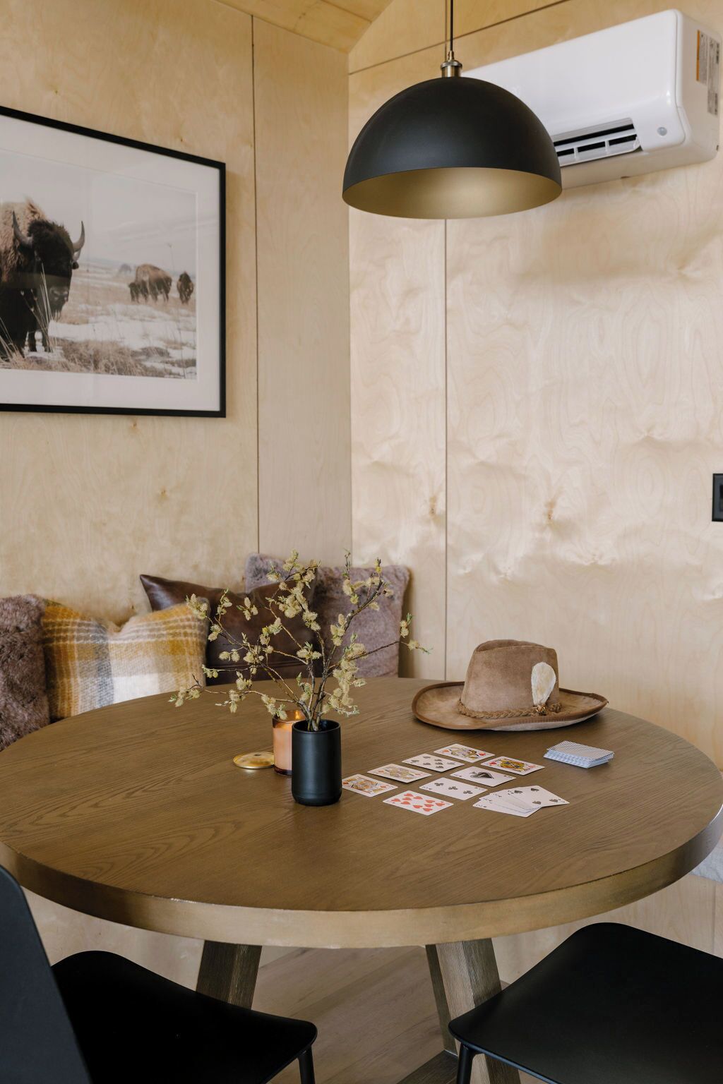 Une table ronde en bois, surmontée d’un luminaire suspendu noir, avec un vase contenant des branches, un jeu de cartes et un chapeau posés sur la table.