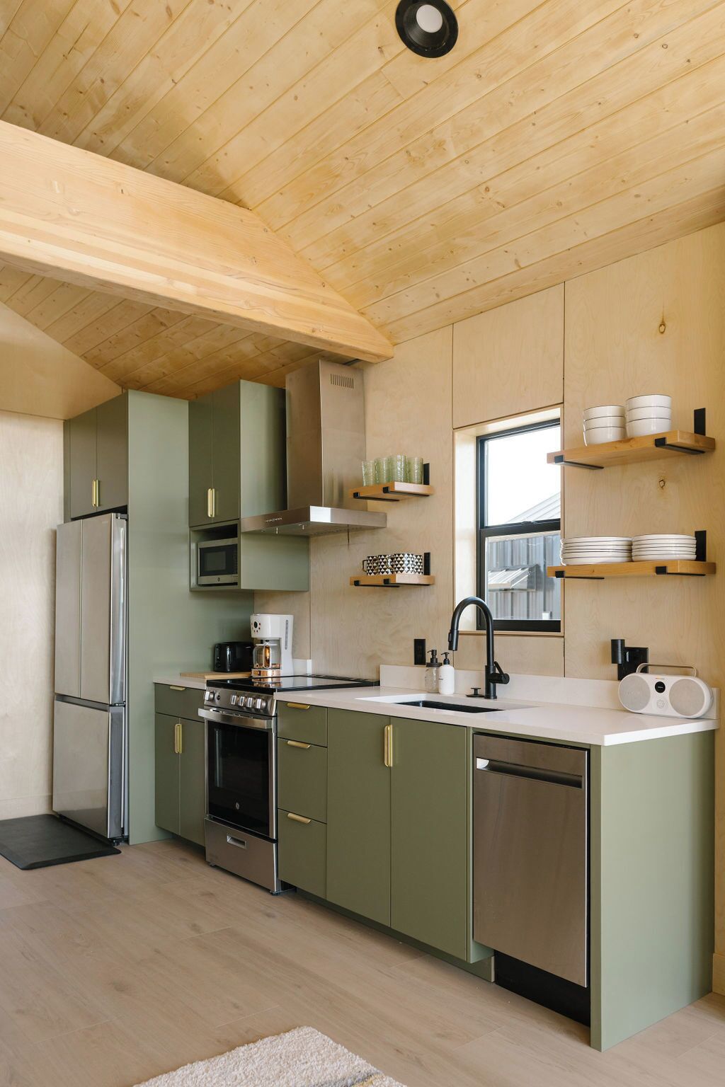 A modern kitchen with wooden ceiling beams, green cabinets, stainless steel appliances, and a window providing natural light.