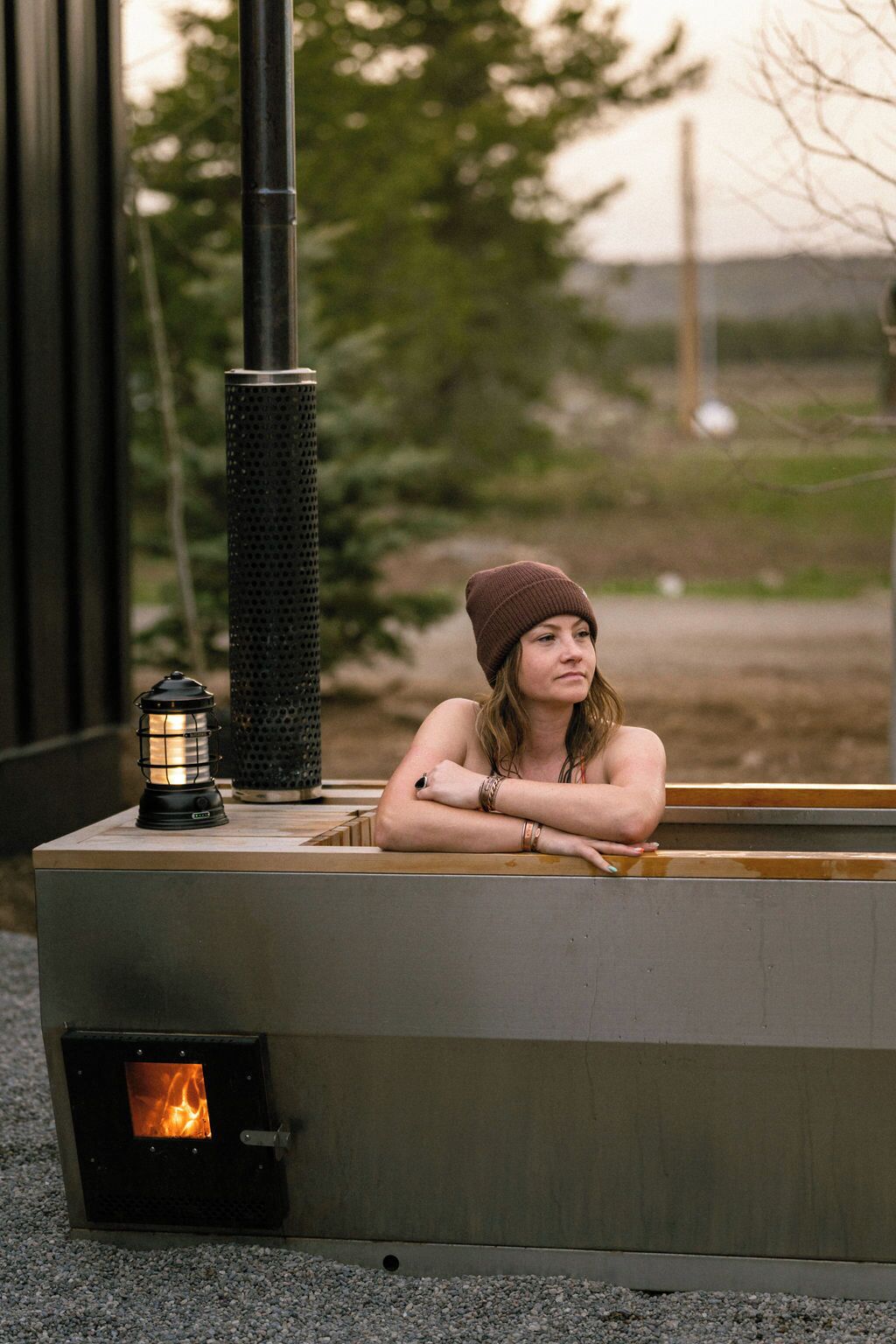 A person leaning on a modern outdoor heater with a visible fire, set against a natural backdrop.