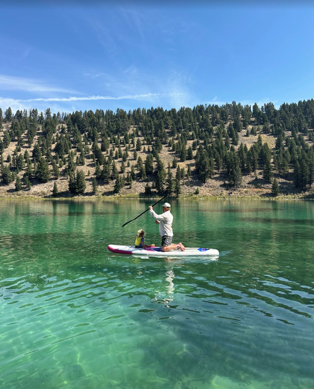 Eine Person steht auf einem Stand-Up-Paddleboard auf einem klaren See, im Hintergrund ein bewaldeter Hügel.