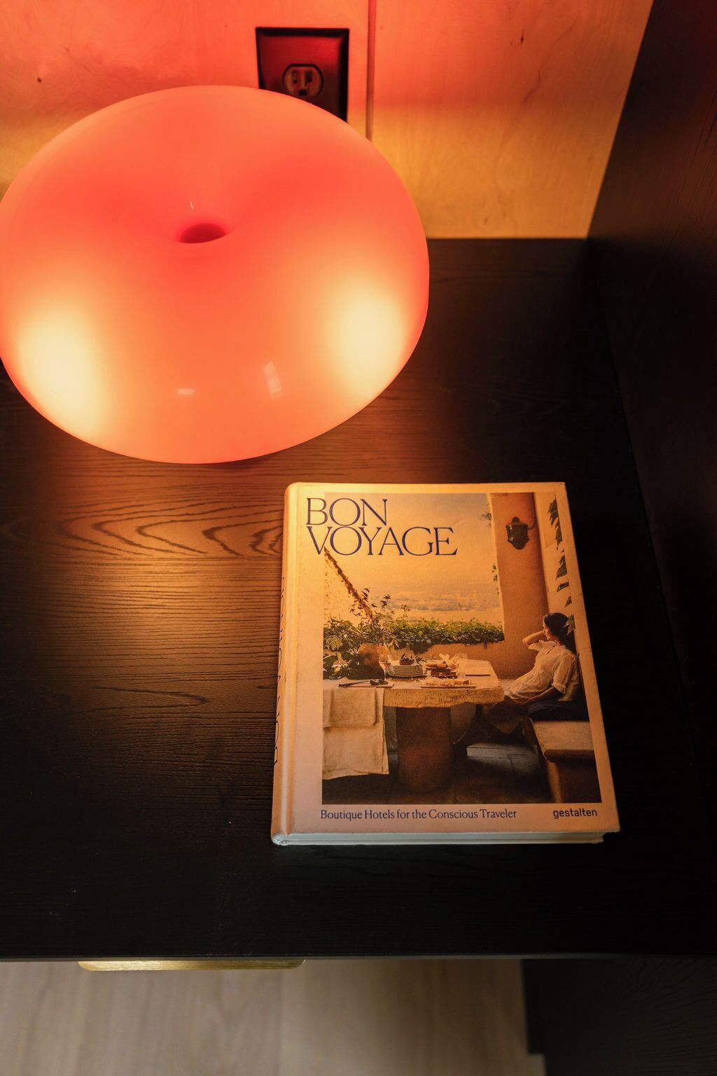 Un livre intitulé « BON VOYAGE » est posé sur une surface en bois, et un luminaire rond est suspendu au-dessus.