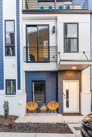 A modern two-story townhouse with a balcony, large windows, and a small outdoor seating area with chairs.