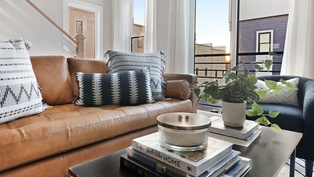 Un salon moderne avec un canapé en cuir marron, des coussins à motifs, une table basse avec des livres et une plante, et une vue sur les bâtiments à travers de grandes fenêtres.