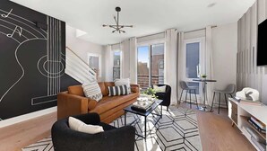Un salon moderne avec un canapé marron, un fauteuil noir et une table basse. On y trouve une œuvre d’art abstraite en noir et blanc accrochée au mur, un lustre et une grande fenêtre avec des rideaux.