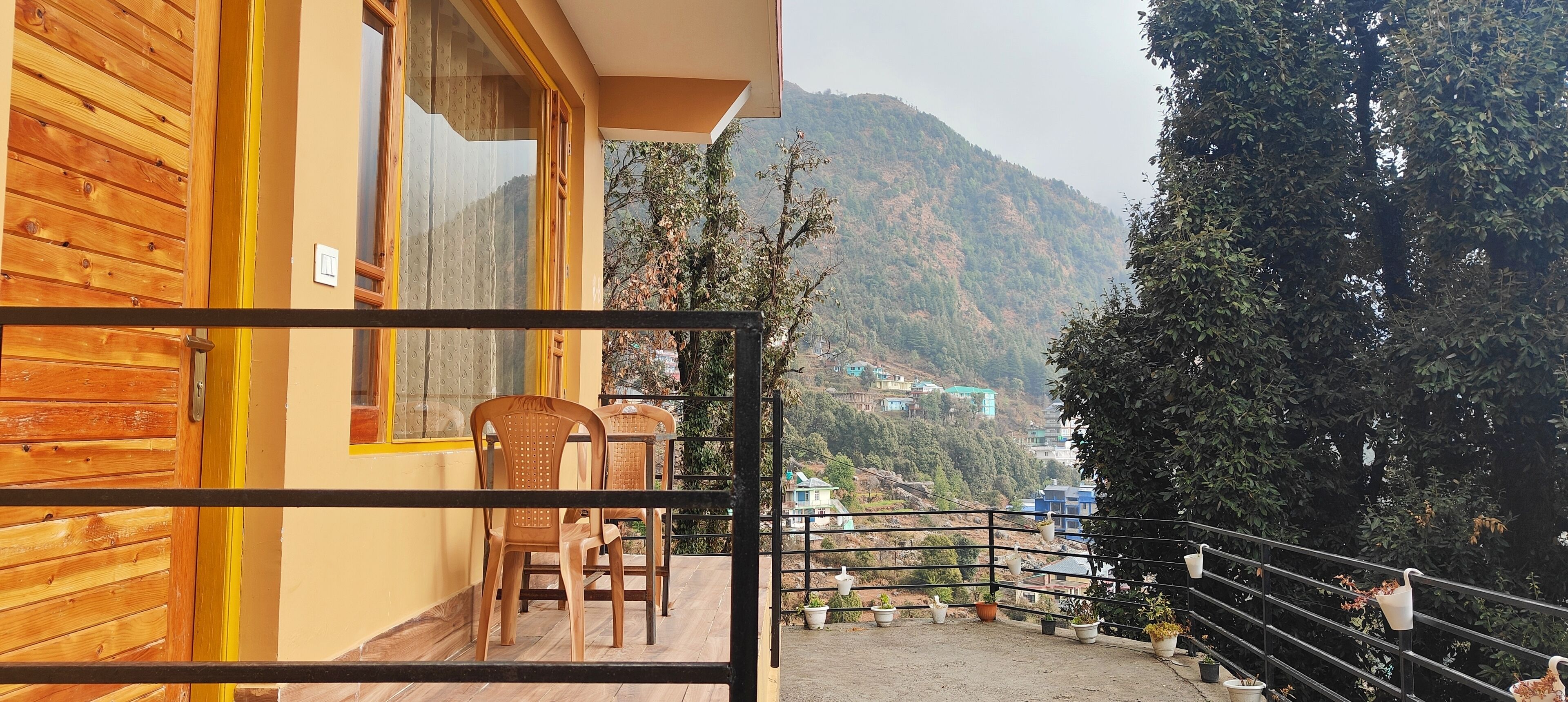 Un balcón con sillas de madera y plantas en macetas, con vistas a un paisaje montañoso y edificios a lo lejos.
