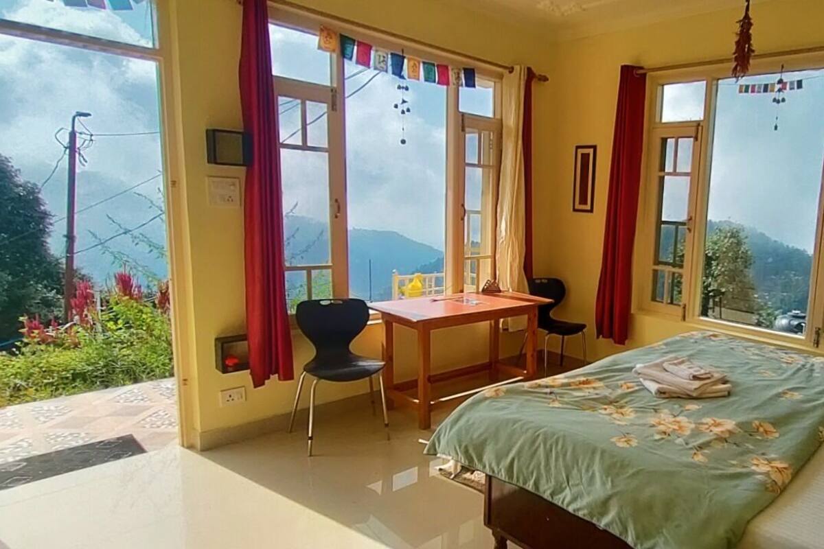 Un dormitorio con cama, escritorio y silla. Amplias ventanas con cortinas, y vista a zonas verdes.