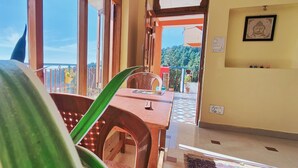 A room with a wooden table, a chair, and a view of a balcony with plants and a railing.